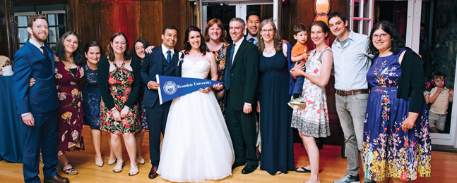 In a wood-paneled room, a group of people pose around a bride and groom holding a Brandeis pennant.